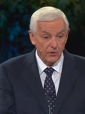 A medium shot of a well-dressed man speaking into a microphone. He is wearing a dark suit, a light-colored shirt, and a patterned tie. The background is out of focus, suggesting a studio or stage setting. His expression is serious.