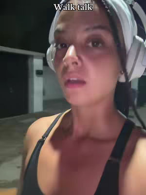 A woman is shown in a selfie-style video, filmed at night with a dark background. She appears to be outdoors, possibly walking. Her hair is pulled back with a white headband, and she is wearing large, white headphones over her ears. She is wearing a black athletic top. Above her head is a text overlay that reads 'Walk talk'.
