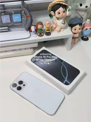 The image is a top-down view of a white desk setting. On the desk, a white iPhone 16 Pro Max is shown next to its white box. Overlayed on the phone box is the text "unboxing iPhone 16 Pro Max White Titanium". Various decorative figurines are placed on a white shelf in the background. The overall composition is clean and bright.