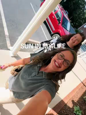 The cover image features a selfie taken from a slightly low angle. The foreground is dominated by a woman close to the camera, wearing glasses and smiling. A second woman is visible behind her, also smiling. A red car and part of a modern building are visible in the background. The image is well-lit, with a casual and happy feel.