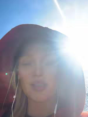 A bright, sunny shot with a woman partially visible. The sun is a prominent element, creating strong backlighting and lens flare. The woman, wearing a dark hat against the sun, is the focus, appearing to be speaking. She is wearing white earbud headphones against a clear blue sky