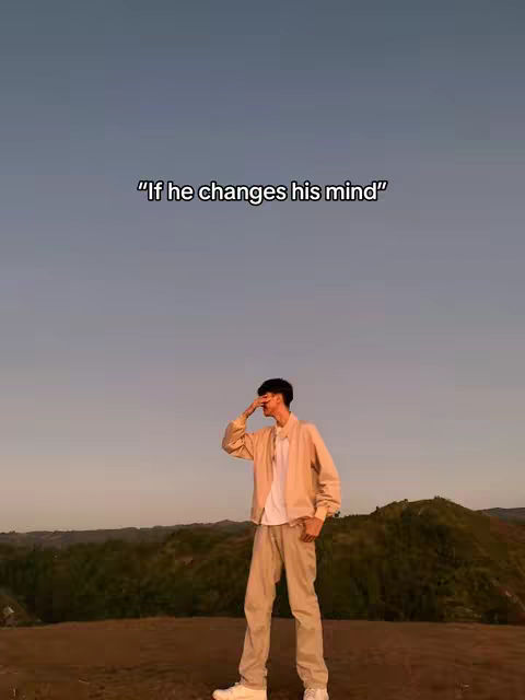 A young man stands on a hilltop, the sky behind him fading from pink to blue. He's wearing a light beige jacket, a white shirt, and tan pants. He's holding his hand up to his face. Text is overlaid at the top of the image.