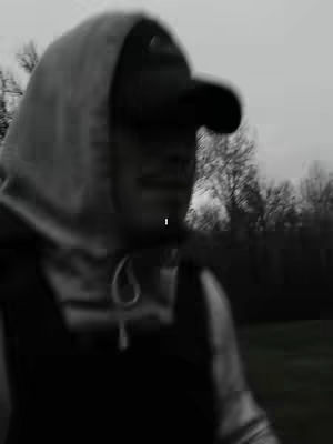 An out-of-focus, grayscale image of a person wearing a hooded sweatshirt and a baseball cap. The person is partially obscured, with the background suggesting an outdoor setting with trees. The image has a blurred, grainy quality.