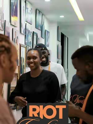 The cover image from a low angle, groups of people in a room. In the center is a dark-skinned woman in a black shirt. The woman is smiling looking slightly to the side. Behind her is a taller man with a baseball cap. The sign with the words "RIOT Conference" is at the bottom. The room is brightly lit.