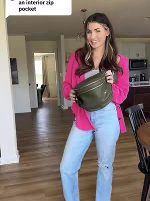 A woman is posing holding a green bag, dressed in a pink jacket, light blue jeans with cut out knees, and a white top. She is inside a home with a wooden floor and light off-white walls. The lighting in the image is natural and soft, enhancing the warm tones throughout the scene. The background elements include an open doorway, which provides a sense of depth. Beyond the doorway, the kitchen island is visible.