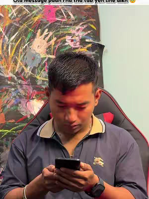 A young man is seated in a black and red gaming chair, looking down at his phone. The background features an abstract painting with a colorful, hand-like design. The man is wearing a dark blue polo shirt, and has a watch on his wrist. The lighting is warm, centered on the man's face and the phone.