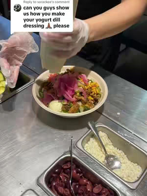 A person in white gloves is pouring a white creamy dressing over a bowl of food. The bowl contains various colorful ingredients. Beside it there are containers with other ingredients.