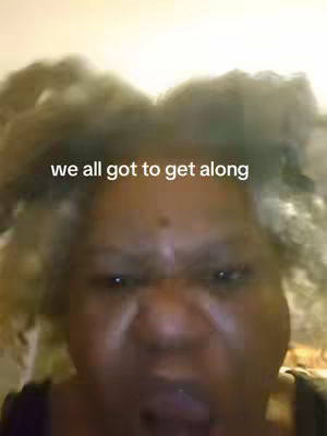 A close-up shot of a woman with short, frizzy, graying hair, looking directly at the camera with a serious expression. The text overlay reads, "we all got to get along". The background is blurred and indistinct.