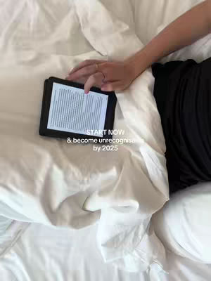 A top-down shot shows a person's hand with a ring resting on a black tablet, which is lying on a bed covered with white sheets. Text overlayed on the image reads 'START NOW & become unrecognisable by 2025'.
