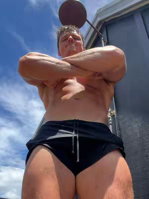 A man positioned beneath an outdoor shower head, shirtless with arms crossed, wearing black shorts. The background is a clear blue sky.