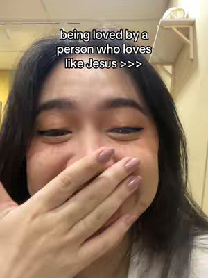 A close-up shot of a young woman covering her mouth with her hand, eyes wide with emotion. The background is blurred, but shows a room with a beige wall and a small wooden shelf. The text overlay says "being loved by a person who loves like Jesus >>>".
