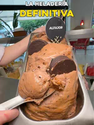 The cover image shows a close-up view of a tray of ice cream. The ice cream itself is light brown with dark chocolate pieces, and what appear to be Alfajor cookies are visible on top. The tray is stainless steel. On the top of the image is the text that reads "LA HELADERÍA DEFINITIVA" in a yellow box with a black border. There is a black and white Alfajor sign sticking out of the ice cream.