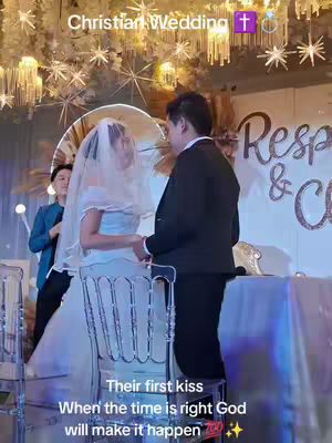 A wedding photo showing a bride and groom holding hands, facing each other. The bride wears a white veil and dress, the groom a black suit. The background is decorated with lights and floral arrangements. Text overlays are present on the image.