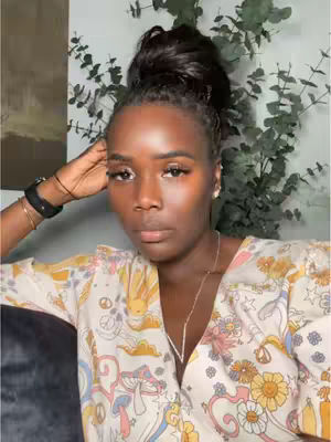A close-up shot features a Black woman with an updo hairstyle, looking directly at the camera. She has full lips, well-defined eyebrows, and long eyelashes with makeup, and also a necklace. She wears a patterned top with various colors and is resting her head and her arm is on a dark surface. The background includes leafy green plants and a partial view of a painting.