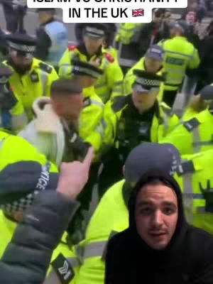 The cover photo shows a scene in the UK with a large group of police officers in uniform, some appearing to interact with people. In the bottom right corner, a person with a dark hoodie is visible in selfie mode, looking at the camera. The background shows a street scene with buildings.