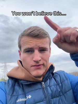 A young man is centered in the frame, looking toward the camera with a serious expression. He has short, light-colored hair and his hand is raised, index finger pointing upwards. The background shows a cloudy sky and some power lines or structures. The man is wearing a blue vest over a light-brown hoodie.