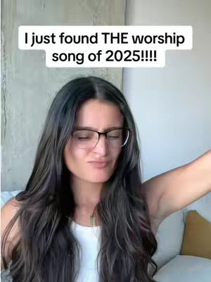 A woman with long dark hair and glasses is in the center of the frame, with a white text box at the top stating "I just found THE worship song of 2025!!!!". She is wearing a white top and the background appears to be a light-colored wall and a portion of a couch.