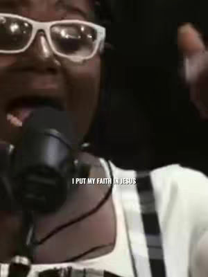 A close-up shot of a person wearing white-framed glasses, a white shirt or top, and a microphone. The person's mouth is open as if they are singing or speaking. The text overlay says, 'I put my faith in Jesus'. The background is dark, obscuring further details.
