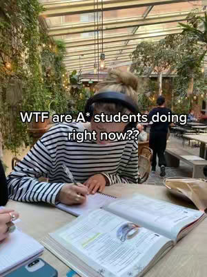 The image shows a woman in a striped shirt with headphones on, seated at a table. She is studying, writing on a pad while several books are open. The table also has a phone and a pink notebook. The background is a bright, indoor garden setting, with lush greenery, and architectural details suggesting a cafe or conservatory.
