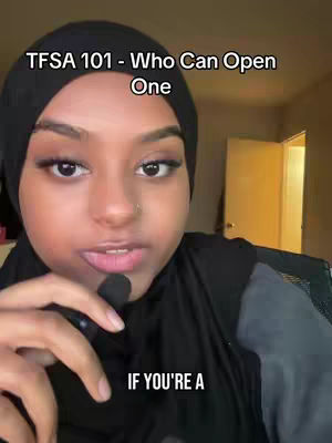 A woman wearing a black hijab is centered in the frame. Above her head is overlaid text in white, and text is also overlaid at the bottom of the frame. A microphone is attached to her collar. The background is blurred, showing a door and other indistinct elements. The lighting seems natural, with a focus on the woman's face.