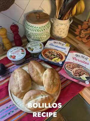 The image showcases a well-lit, overhead shot of a table setting focused on food preparation. A round, woven plate holds several golden-brown bolillos, or bread rolls, arranged in a semi-circle. Surrounding the bread are several elements: a package of Del Real Barbacoa and Carnitas, containers of salsa and crema, and various kitchen utensils. The scene is set on a vibrant, patterned textile. The overall composition is warm and inviting, emphasizing a culinary theme.