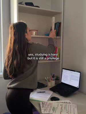 A young woman with long brown hair is standing in front of a white bookshelf, reaching for a book. She is wearing a gray sweater and black leggings. A desk with a laptop and papers is in the foreground. Text on the image reads 'yes, studying is hard but it is still a privilege.'