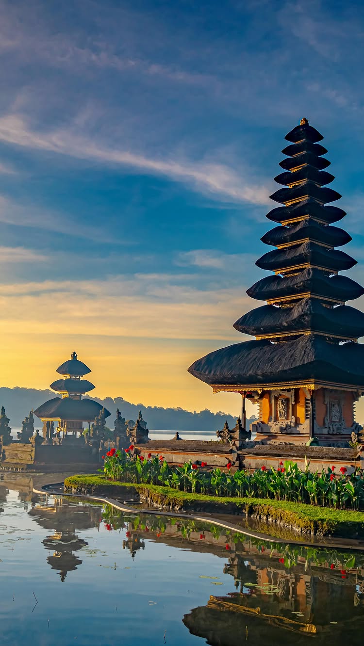A serene landscape features a tiered pagoda reflected in tranquil water. The sky transitions from a golden sunrise to a clear blue. Lush greenery and vibrant red flowers flank the water's edge.