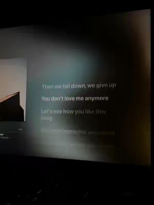 The image shows a screen displaying lyrics in white text against a blurred, dark background. The left side of the screen shows a part of an image. The overall composition focuses on the lyrics from the song.