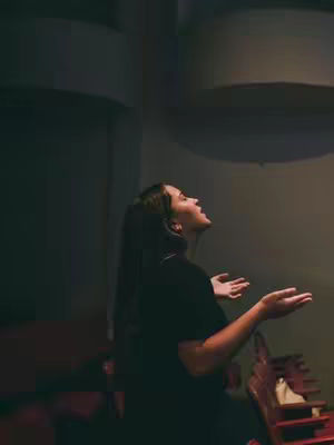 A woman with long hair stands in profile, facing upwards with arms outstretched and palms up, in a gesture of prayer or supplication. The background is dark, suggesting a dimly lit auditorium or performance space. The lighting highlights the woman's face and form, creating a sense of reverence.