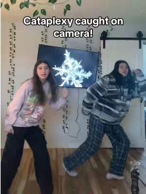 Two young women are dancing in a room. The background includes a TV showing a snowflake pattern, and there are plants. The hook shows the words "Cataplexy caught on camera!"