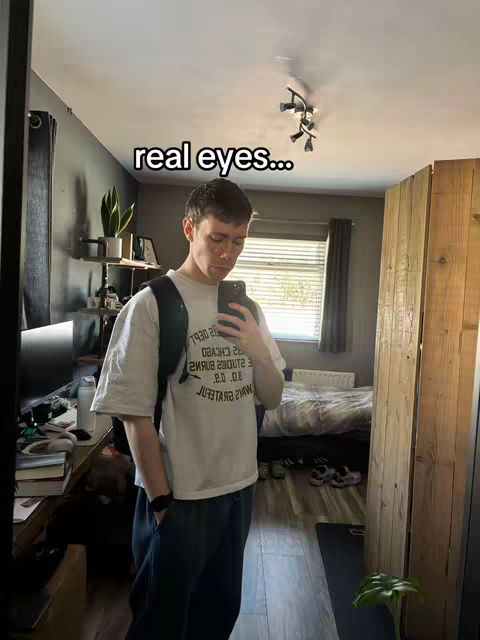 A mirror selfie shows a young man in a room. The man is casually dressed in a white t-shirt and dark pants, with a backpack slung over one shoulder. He's holding a phone up to his face, appearing to take a photo. The room has a bed, a window with blinds, and some decor visible in the background. The text overlay says "real eyes..."