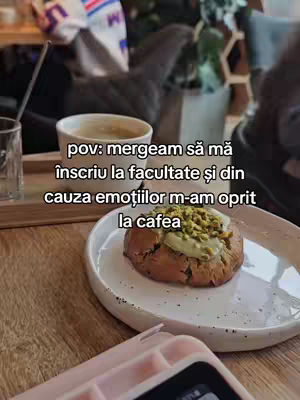 The image shows a close-up of a cafe setting. A small white coffee cup is visible on a wooden surface. Beside it, a glass and a plate with pastry can be seen. In the background, are elements indicating a cafe or restaurant, like the light and the decoration, and some blurred objects.
