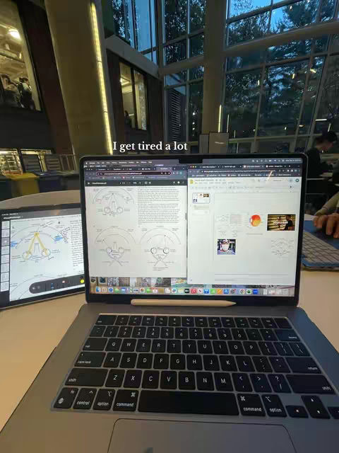 A laptop sitting on a white desk. The laptop is open, displaying what appears to be academic work. A pencil rests on the keyboard. To the left is a tablet showing diagrams. Additionally, the image captures a bright, spacious setting, possibly a library or study area, with large windows and natural light.