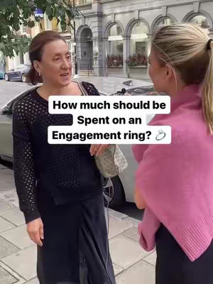 Two women are engaged in a street interview. The woman on the left, the interviewer, is wearing a dark top and black skirt, holding a purse, and gesturing with her hands. The woman on the right is wearing a pink sweater. The background is a well-lit street scene with a building.