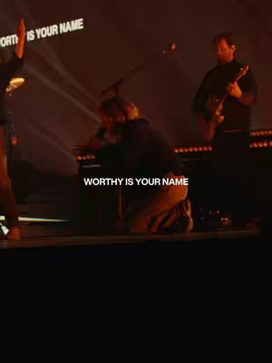 The cover image depicts a stage performance with a band. A person is kneeling, possibly in prayer or worship, with their hands clasped. Text overlayed reads, 'WORTHY IS YOUR NAME.' The background is dark with lighting focused on the performers.