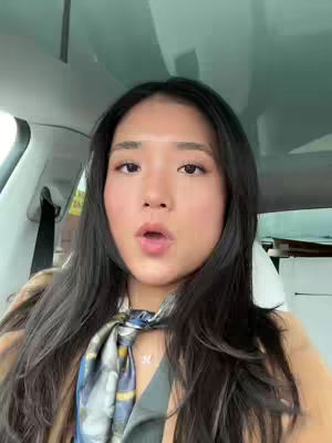 A woman with long black hair is seated in a car, looking directly at the camera with a slightly surprised expression. She is wearing a scarf with a blue and gray pattern. The car's interior is visible, with the headliner and seat visible in the background.