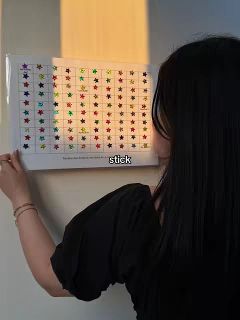 A person with long black hair is holding an opened book that is filled with colorful star-shaped stickers. The person is seen from the back, with their face partially visible on the right side of the frame. The background consists of a white wall and a patch of golden sunlight.