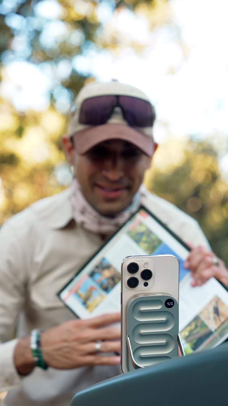 A man with a cap and sunglasses, holding a phone with a power bank attached, and a book, with a blurred background of a natural setting.