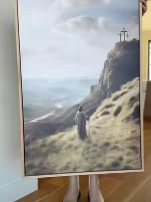 A painting held by a pair of legs, showing a solitary figure walking towards three crosses on a hill, set against a landscape with a cloudy sky.