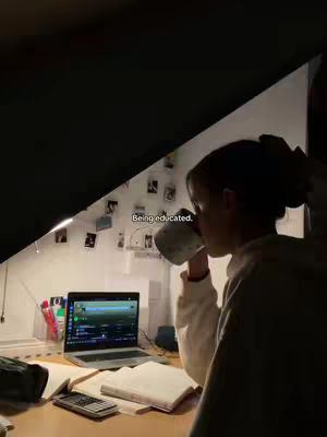 A dimly lit room shows a person, viewed from the side, drinking from a mug. The person is in the foreground, with a desk, laptop, and items like books and a calculator visible in the middle ground. In the background, some photos are taped to the wall near a light.