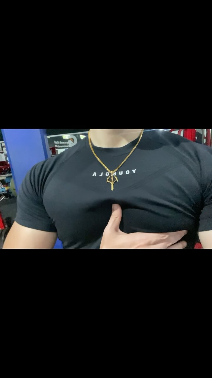 A man in a black t-shirt touches his chest. He is wearing a gold necklace and is in a gym setting. The background is blurry, suggesting a workout environment.