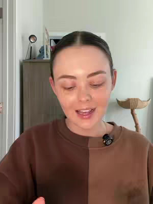 A woman with light skin and brown hair is centered in the frame, looking at the camera and speaking. She is wearing a two-toned sweater, half brown and half dark brown. The background shows a room with cream-colored walls, a light-colored cabinet, and a piece of furniture on the right. A microphone is attached on her sweater.