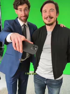 Two men are standing in front of a green screen. The man on the left is wearing a blue suit, holding a phone, and smiling. The man on the right is wearing a black bomber jacket. Both men have facial hair and appear to be in their 30s or 40s.