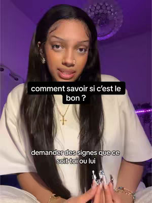 A young woman with long dark hair and a white top is centered in the frame. She is looking at the camera and has text overlayed on the image. The background is a blurry room with purple lighting. She has multiple pieces of jewelry on and appears to be indoors.