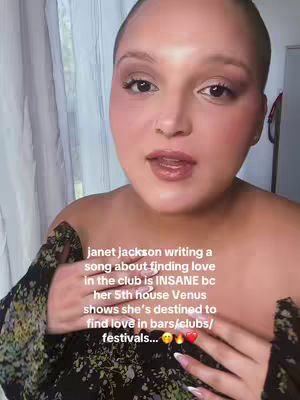 A woman with light skin leans forward in a selfie-style video, speaking directly to the camera. She appears to be indoors, with a soft, blurred background suggesting a room with a curtain. The woman has a shaved head, and she is wearing makeup, including blush and lip color. Her expression is engaged, and her hands are visible, one near her neck with long, light pink nails, and the other near her chest touching a floral patterned top. Text overlay, which is added to the image, wraps around the form of the front of her body, creating a frame for the content.