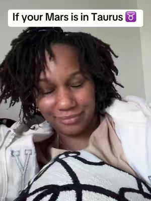 A person with dark, tightly curled hair in a natural style sits with closed eyes, looking slightly to the side and smiling gently. They are wearing a white, open jacket over a light-colored top. The background appears to be neutral, and they are holding a pillow or cushion with a black and white geometric pattern in their lap.