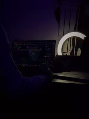The cover image shows a dimly lit scene with a person working on a laptop at a desk. The screen displays a graph, indicating a financial or trading context. A ring light is positioned to the right, providing ambient lighting. The setting suggests a focused, nighttime work environment.