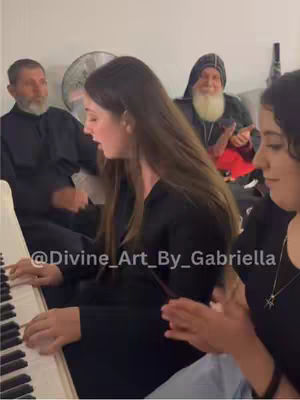 A well-lit indoor scene showing a woman playing the piano, with two religious figures in the background. The woman is wearing a black top, and the setting appears to be a living room with plain walls.