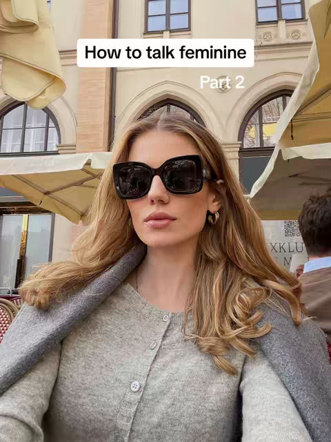 A woman with long blonde hair is wearing a grey sweater and large black sunglasses, looking directly at the camera. The background shows a blurred outdoor setting with building facades, awnings, and hints of other people. Text overlay reads 'How to talk feminine Part 2.'