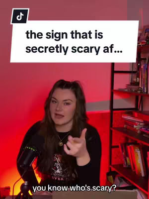 A woman with long brown hair is featured against a red-lit background in the video cover image. She points towards the viewer with her right hand, while a microphone is positioned to her left. Text overlays in white rectangular boxes, reading "the sign that is secretly scary af..." and "you know who's scary?" appear at the top and bottom of the frame. The overall composition is well-lit and appears to be a vlog-style recording.